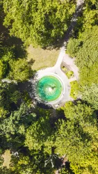 Ein runder Teich umgeben von dichtem Grün. Die Aufnahme zeigt eine klare Wasseroberfläche und gepflegte Gartenanlagen.