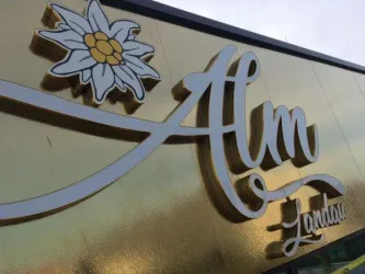 Ein goldenes Schild mit dem Schriftzug "Alm" und einer Edelweiss-Blume. Es zeigt eine elegante und einladende Präsentation.