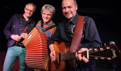 Drei Musiker stehen auf der Bühne und spielen verschiedene Instrumente. Einer hat eine Gitarre, während ein anderer ein Akkordeon hält.