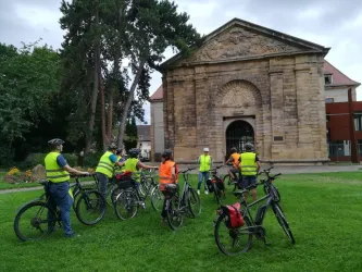 Eine Gruppe von Radfahrern in leuchtenden Westen steht auf einer Wiese vor einem historischen Gebäude. Im Hintergrund sind Bäume und ein bewölkter Himmel sichtbar.