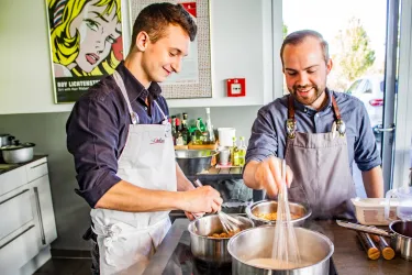 Zwei Männer kochen in einer modernen Küche. Beide tragen Schürzen und arbeiten an verschiedenen Töpfen.