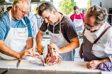 Drei Männer in Schürzen schneiden gemeinsam Fleisch in einer modernen Küche. Im Hintergrund sind weitere Teilnehmer eines Kochkurses zu sehen.
