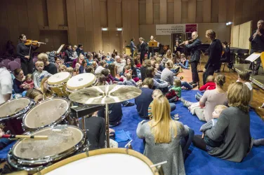 Eine Gruppe von Kindern und Erwachsenen sitzt auf dem Boden und hört einer musikalischen Darbietung zu. Im Vordergrund sind Schlaginstrumente zu sehen, während der Raum voller Musik und Freude ist.