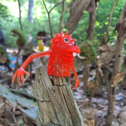 Eine kleine rote Plastikfigur mit spitzen Ausstülpungen steht auf einem Stück Holz. Im Hintergrund sind Waldboden und grüne Pflanzen sichtbar.