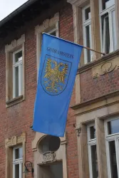 Eine blaue Fahne mit dem Wappen von Godramstein hängt neben einem historischen Gebäude. Im Hintergrund sind Fenster und rote Ziegelmauern zu sehen.