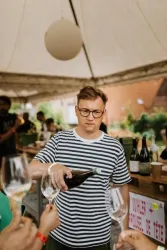 Eine Person mit Brille gießt Wein in ein Glas. Im Hintergrund sind weitere Menschen und eine festliche Atmosphäre zu sehen.