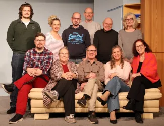 Eine Gruppe von Menschen sitzt auf einem Sofa und lächelt in die Kamera. Sie scheinen Teil eines Theater- oder Filmprojekts zu sein.