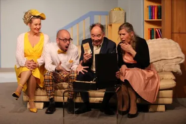 Vier Personen sitzen auf einem Sofa und schauen überrascht auf einen Koffer. Sie sind in bunten Kostümen gekleidet und scheinen in einer humorvollen Szene zu sein.