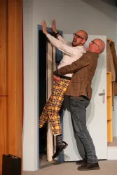 Zwei Männer in komischen Outfits stehen im Türrahmen. Einer hält den anderen auf humorvolle Weise hoch.