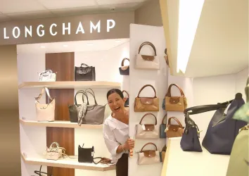 Eine fröhliche Frau steht in einem Modegeschäft und betrachtet Handtaschen. Im Hintergrund sind die Taschen von Longchamp auf Regalen ausgestellt.