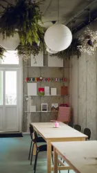 Ein helles, modernes Atelier mit einem großen Tisch und Stühlen. An der Wand hängen kreative Materialien und Pflanzen im Raum sind dekorativ angeordnet.