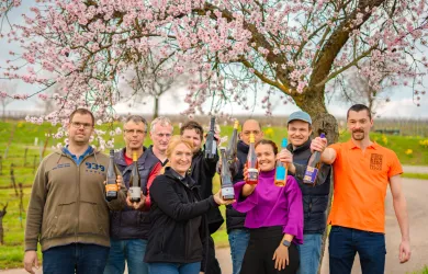 Eine Gruppe von Menschen steht unter einem blühenden Baum und hält verschiedene Getränke in der Hand. Im Hintergrund sind Weinberge und eine Frühlingslandschaft zu sehen.