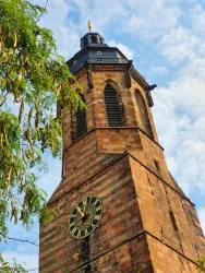 Ein historischer Turm mit einer großen Uhr und gotischen Fenstern. Der Himmel ist teilweise bewölkt und Bäume sind im Vordergrund sichtbar.