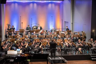 Ein Orchester spielt auf einer Bühne mit bunten Lichtern im Hintergrund. Der Dirigent leitet die Musiker, die verschiedene Instrumente spielen.