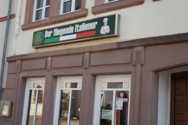 Das Bild zeigt das Restaurant "Der fliegende Italiener" mit einem auffälligen Schild. Die Fassade des Gebäudes ist in einem hellen Farbton gehalten und hat große Fenster.