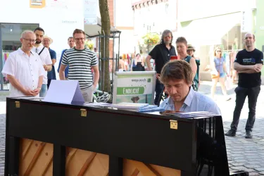 Ein Musiker spielt Klavier auf der Straße, während eine kleine Menge zuschaut. Es ist ein sonniger Tag mit einer lebhaften Atmosphäre.