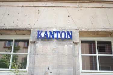 Das Bild zeigt ein Schild mit der Aufschrift "KANTON" an einer wand mit Fenster. Der Hintergrund besteht aus beige-farbenem Mauerwerk.