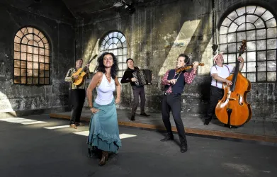 Vier Frauen in eleganten Kleidern sitzen um ein Klavier. Sie lächeln und posieren zusammen für das Foto.