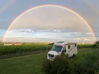 Ein Wohnmobil steht auf einer Wiese unter einem Regenbogen. Die Landschaft ist grün mit Weinfeldern im Hintergrund.