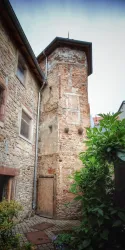 Ein alter, teilweise verfallener Turm aus Stein steht neben einem Gebäude. Umgeben von Pflanzen und einem gepflasterten Weg schafft er eine interessante Atmosphäre im Innenhof.