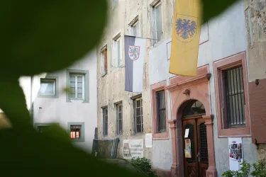 Ein historisches Gebäude mit einer markanten Fassade und Fahnen. Im Vordergrund sind grüne Blätter zu sehen, die einen Teil des Bildes rahmen.