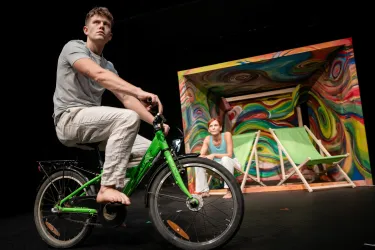 Ein junger Mann sitzt barfuß auf einem grünen Fahrrad, während eine Frau im Hintergrund auf einem Stuhl sitzt. Die Kulisse zeigt bunte kreative Elemente, die eine lebendige Szene schaffen.