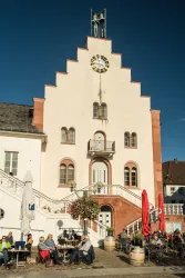 Ein historisches Gebäude mit einem markanten, spitzen Dach und einer Uhr. Vor dem Gebäude sitzen Menschen in einem Café bei schönem Wetter.