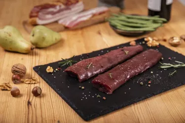Zwei Stücke rotes Fleisch auf einem schwarzen Steinbrett, dekoriert mit Rosmarin. Im Hintergrund sind Birnen, Speck und grüne Bohnen zu sehen.