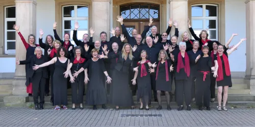 Eine fröhliche Gruppe von Sängern in schwarz-rot posiert vor einem Gebäude. Sie zeigen ihre Begeisterung mit erhobenen Händen und lächeln.