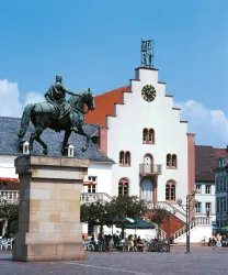 Ein beeindruckendes Reiterstandbild steht vor einem historischen Gebäude mit einer auffälligen Uhr. Die Szene ist lebhaft mit Menschen und Fahrrädern im Hintergrund.