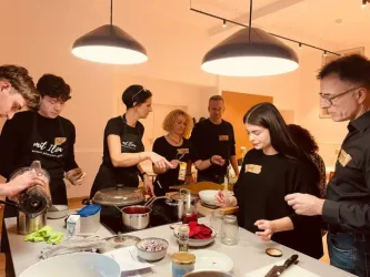 Eine Gruppe von Personen bereitet gemeinsam Essen in einer modernen Küche vor. Alle tragen schwarze Schürzen und arbeiten an verschiedenen Zutaten und Kochgeräten.