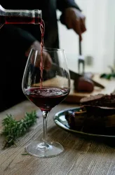 Ein Glas Rotwein wird eingeschenkt, während im Hintergrund ein Stück Fleisch auf einem Brett geschnitten wird. Neben dem Glas liegt frisches Rosmarin.
