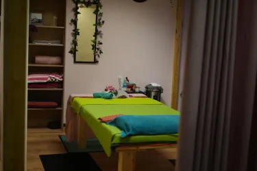 Ein ruhiger Massageraum mit einem grünen Tisch und bunten Handtüchern. Im Hintergrund sind Regale mit weiteren Textilien und einer Spiegelwand zu sehen.