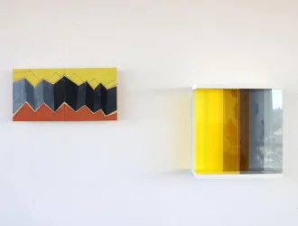 Zwei Kunstwerke an einer Wand. Links ein farbenfrohes Gemälde mit geometrischen Formen und rechts eine transparente Box mit gelben und grauen Farben.