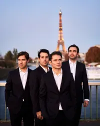 Vier Männer in Anzügen stehen auf einer Brücke in Paris. Im Hintergrund ist der Eiffelturm bei Sonnenuntergang zu sehen.
