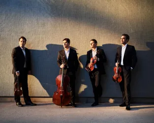 Vier Männer in eleganter Kleidung stehen in einer Reihe. Drei halten Geigen, einer ein Cello in der Hand. Die Sonne (von links) wirft lange Schatten der Männer nach rechts ins Bild. Der Hintergrund ist in grau gehalten.