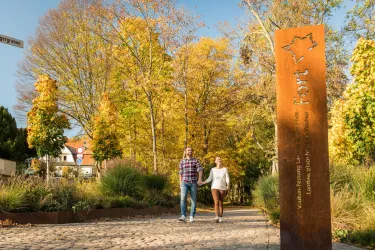 Ein hübscher Park mit buntem Herbstlaub und Spaziergängern. Im Hintergrund steht ein informatives Schild.
