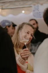 Eine lächelnde Frau hält ein Weinglas in der Hand. Im Hintergrund sind weitere Personen zu sehen.