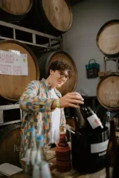 Ein junger Mann mit einer bunten Jacke schenkt Wein aus einer Flasche ein. Im Hintergrund sind Holzfässer und mehrere Flaschen sichtbar.