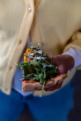 Eine Person hält ein Stück Pizza mit frischem Rucola und Käse. Der Hintergrund ist unscharf, während die Pizza im Vordergrund klar sichtbar ist.
