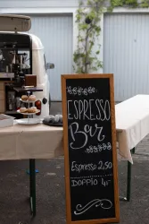 Ein auf einem Tisch stehendes Schild mit der Aufschrift "Espresso Bar". Neben dem Schild sind einige Kuchen und ein Kaffeewagen zu sehen.