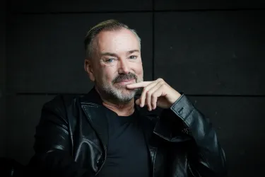 Ein Mann mit einem freundlichen Gesicht sitzt entspannt und lächelt. Er trägt eine schwarze Lederjacke und hat eine nachdenkliche Pose.
