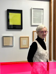 Eine ältere Frau mit weißem Haar und Brille steht vor einer Wand mit verschiedenen gerahmten Kunstwerken. Die Bilder sind in neutralen Farben, darunter ein leuchtend gelbes Werk.