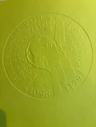 Eine reliefartige Darstellung von Hildegard von Bingen auf gelbem Hintergrund. Die Plakette zeigt ihren Namen und Lebensdaten sowie dekorative Elemente.