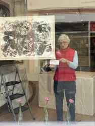 Eine ältere Frau steht vor einem großen Kunstwerk in einem Schaufenster. Auf einem Tisch sind Blumen in Glasflaschen arrangiert.