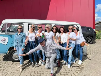 Eine Gruppe von Frauen steht fröhlich vor einem Oldtimer-Bus und posiert für das Foto. Im Hintergrund ist ein rotes Gebäude und ein schwarzes Auto zu sehen.