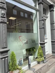Ein Friseursalon mit der Beschriftung "Jessis Hair & Style" im Fenster. Vor dem Salon stehen einige Pflanzen in Töpfen.