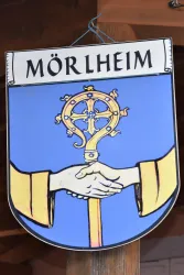 Ein Wappen von Mörlheim, das zwei Hände zeigt, die sich zur Begrüßung reichen. Darüber befindet sich ein goldenes Kreuz und der Name "Mörlheim".
