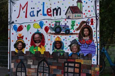 Eine bunte Wandmalerei mit Luftballons, einem Traktor und einer kleinen Häuser-Szene. Fünf Personen stecken mit ihren Köpfen in die Ausschnitte, die wie eine Mauer gestaltet sind.