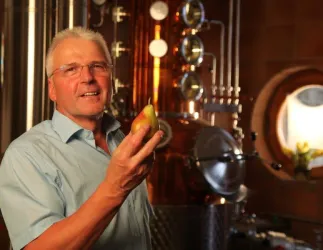 Willi Peter hält eine Birne in der Hand und steht vor einer Brennerei. Im Hintergrund sind glänzende Metallrohre und eine große Fensteröffnung zu sehen.